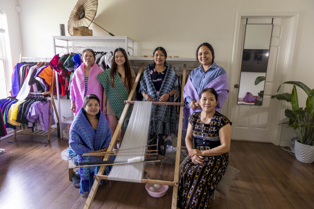 Six women pose around a wooden floor loom inside a studio space. Several wear handwoven textiles draped over their shoulders. Clothing racks and woven garments are visible behind them.
