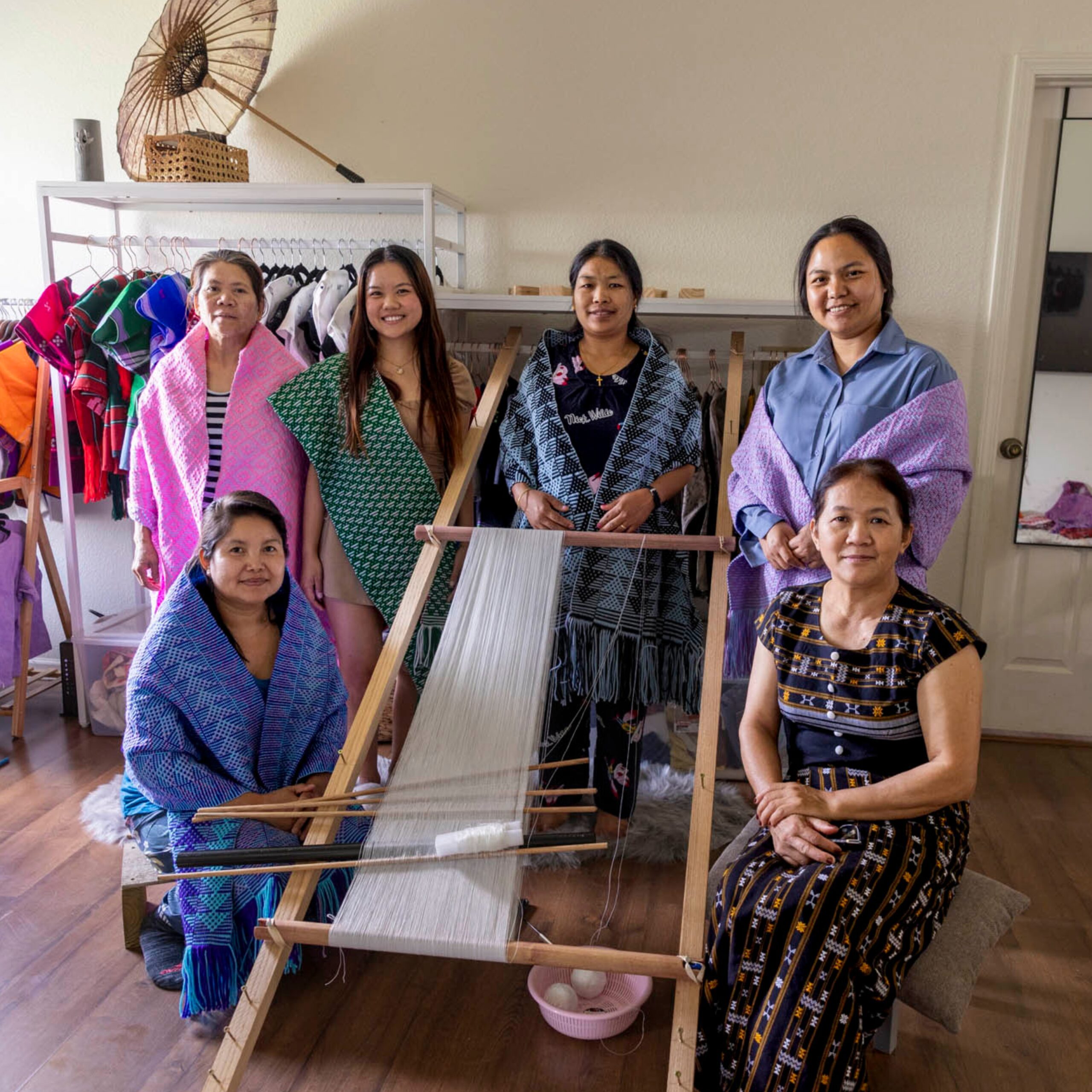 Six women pose around a wooden floor loom inside a studio space. Several wear handwoven textiles draped over their shoulders. Clothing racks and woven garments are visible behind them.