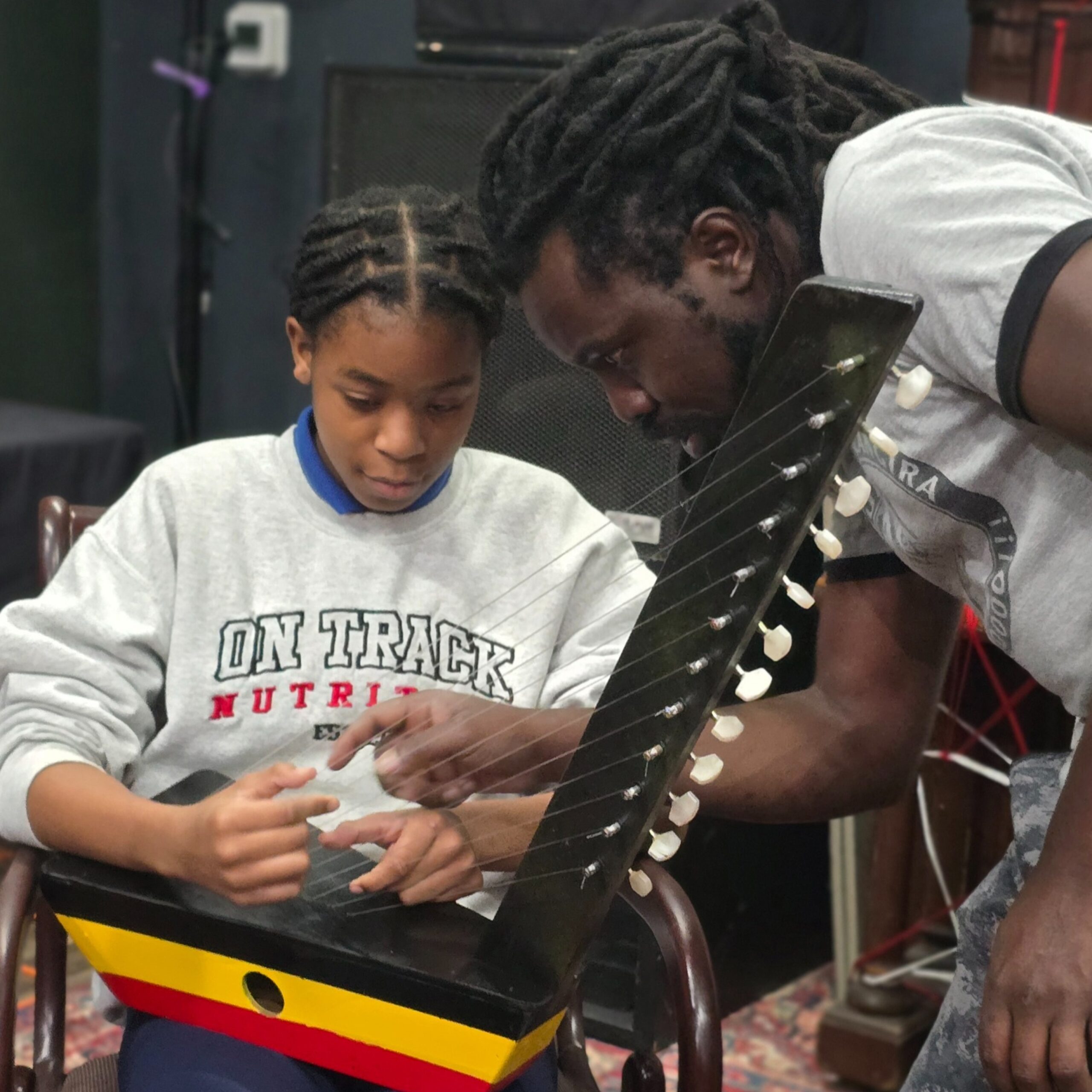 An adult leans in to guide a young person as they pluck a traditional stringed instrument with multiple tuning pegs. Both focus closely on the strings during a music lesson in an indoor community space.