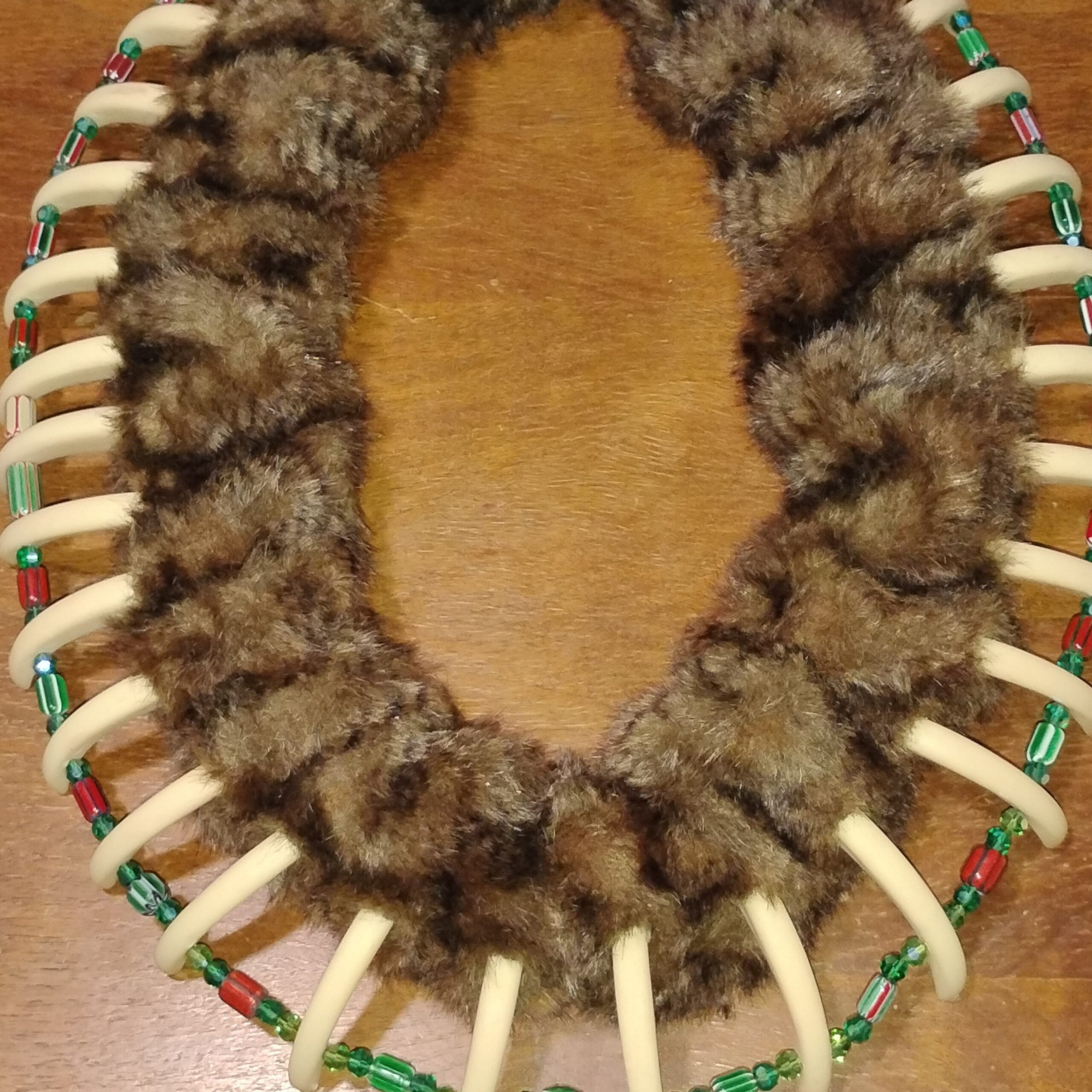 A close-up of a handcrafted necklace made from fur, bone-colored curved pieces, and red and green beads, arranged symmetrically on a wooden surface.