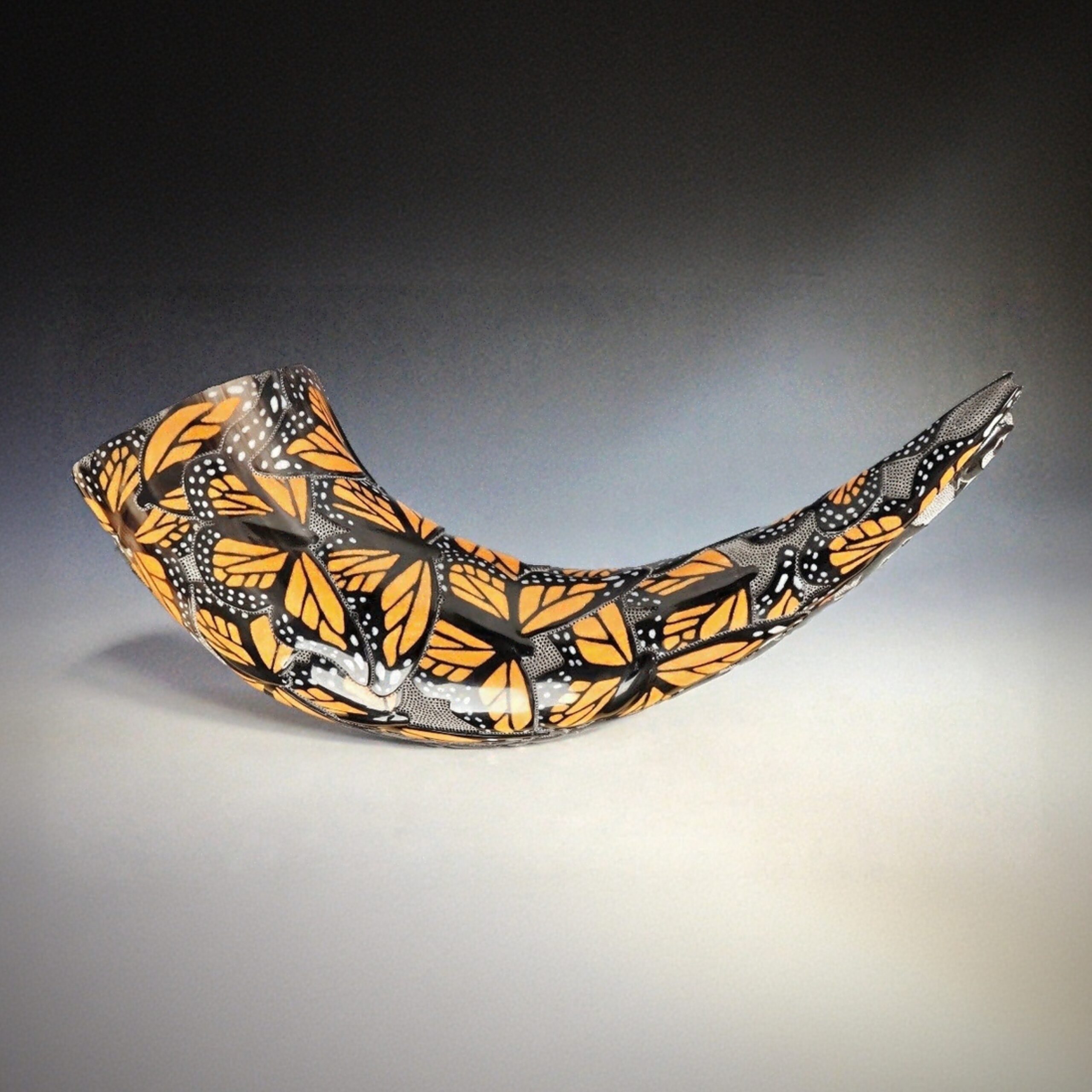 A polished, curved buffalo horn vessel decorated with an intricate monarch butterfly pattern in orange, black, and white rests against a neutral background.