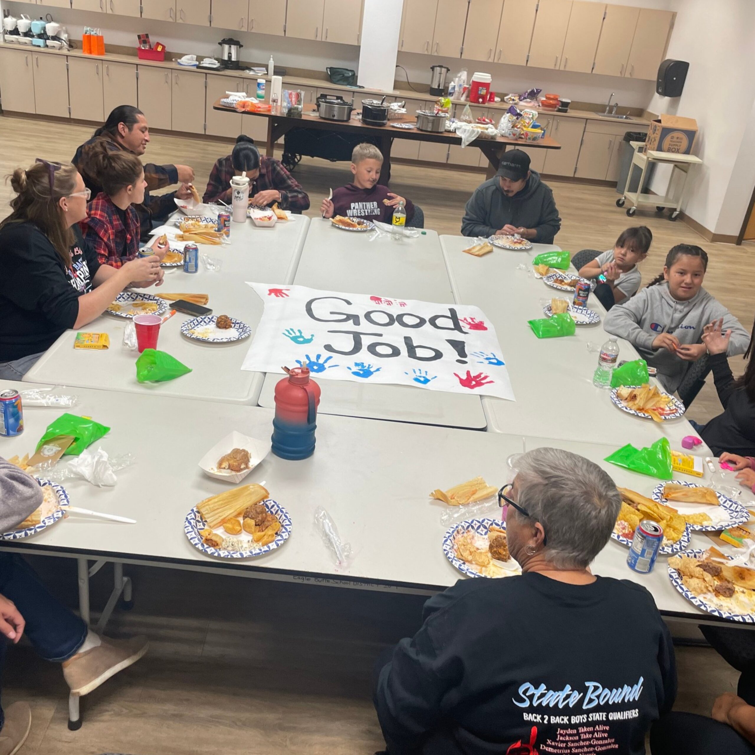 Community members of multiple ages sit around connected tables, sharing a meal during an Keya gathering. A handmade sign reading “Good Job!” with colorful handprints is placed at the center of the tables in a community room kitchen.