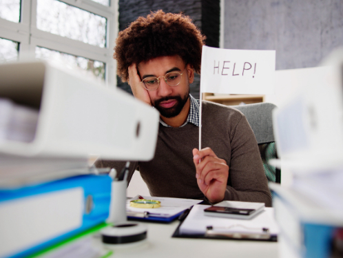 A man with dark skin tone and brown hair, mustache and goatee sits at a desk resting his head on his hand and holding a sign that reads HELP