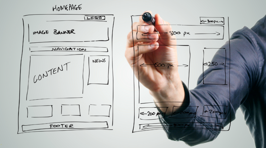 Light colored hand holding black marker drawing a website wireframe against a gray background. The person is wearing a dark colored shirt.