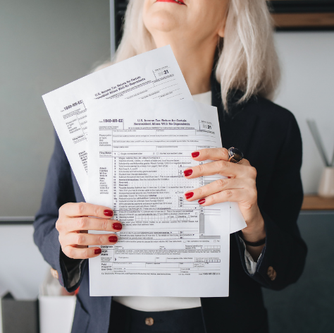 A woman with shoulder length white hair and wearing a black suit looks upward and holds blank tax forms in front of her