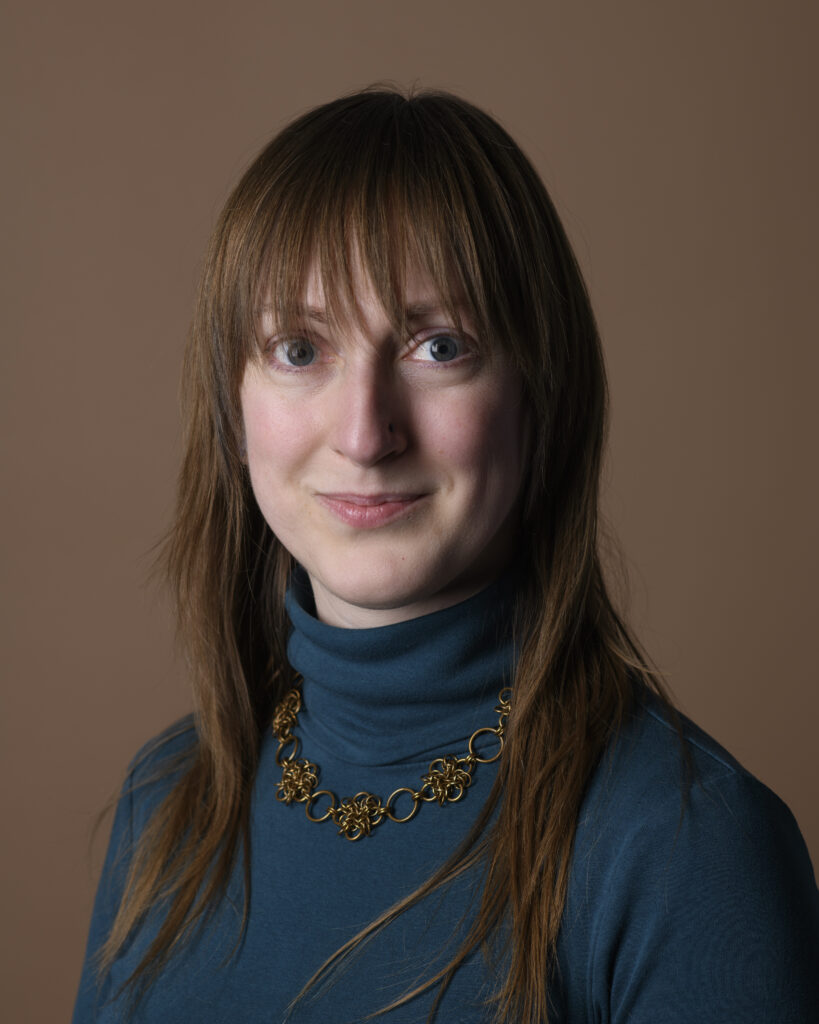 A woman with light skin and medium brown hair with bangs, wearing a deep teal turtleneck and a brown metallic necklace.
