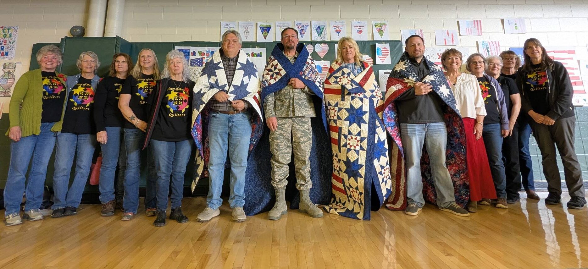 A group of people standing in a line. Four of them have red, blue, and white quilts over their shoulders.