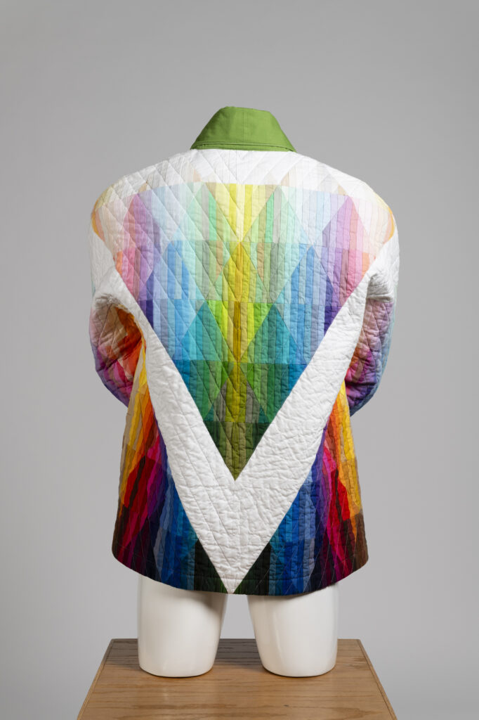 A quilted coat with a multicolored pattern of rainbow patterned triangles and sleeves on a mannequin, facing away.