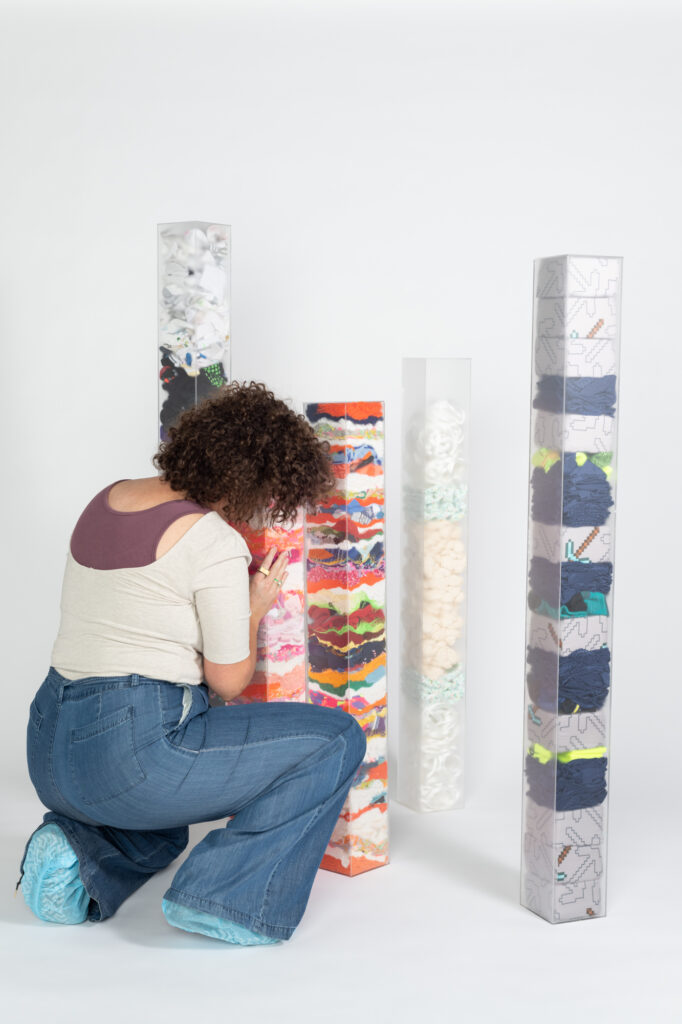 A woman in a white shirt, blue jeans, and blue covers over her shoes kneeling to adjust colorful rectangular sculptures against a white paper backdrop.