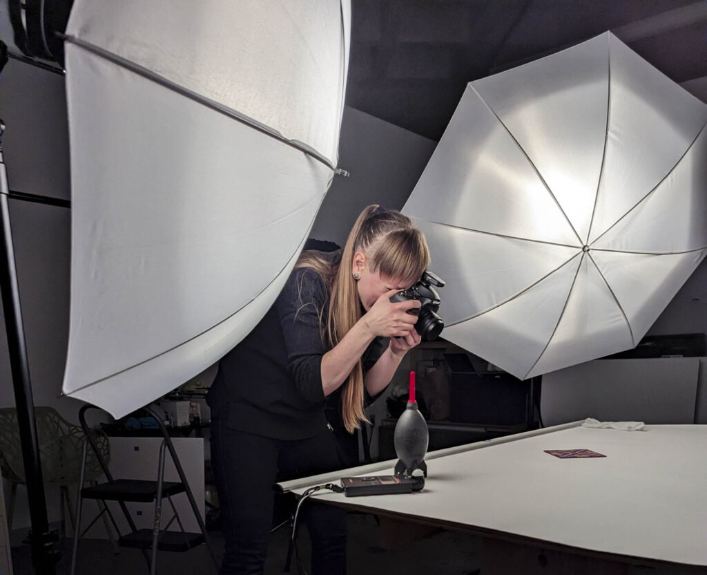 A woman with long light brown hair in a ponytail holding a camera up to her eye, surrounded by lights behind large white umbrellas, as she photographs a piece of art on a flat gray surface.