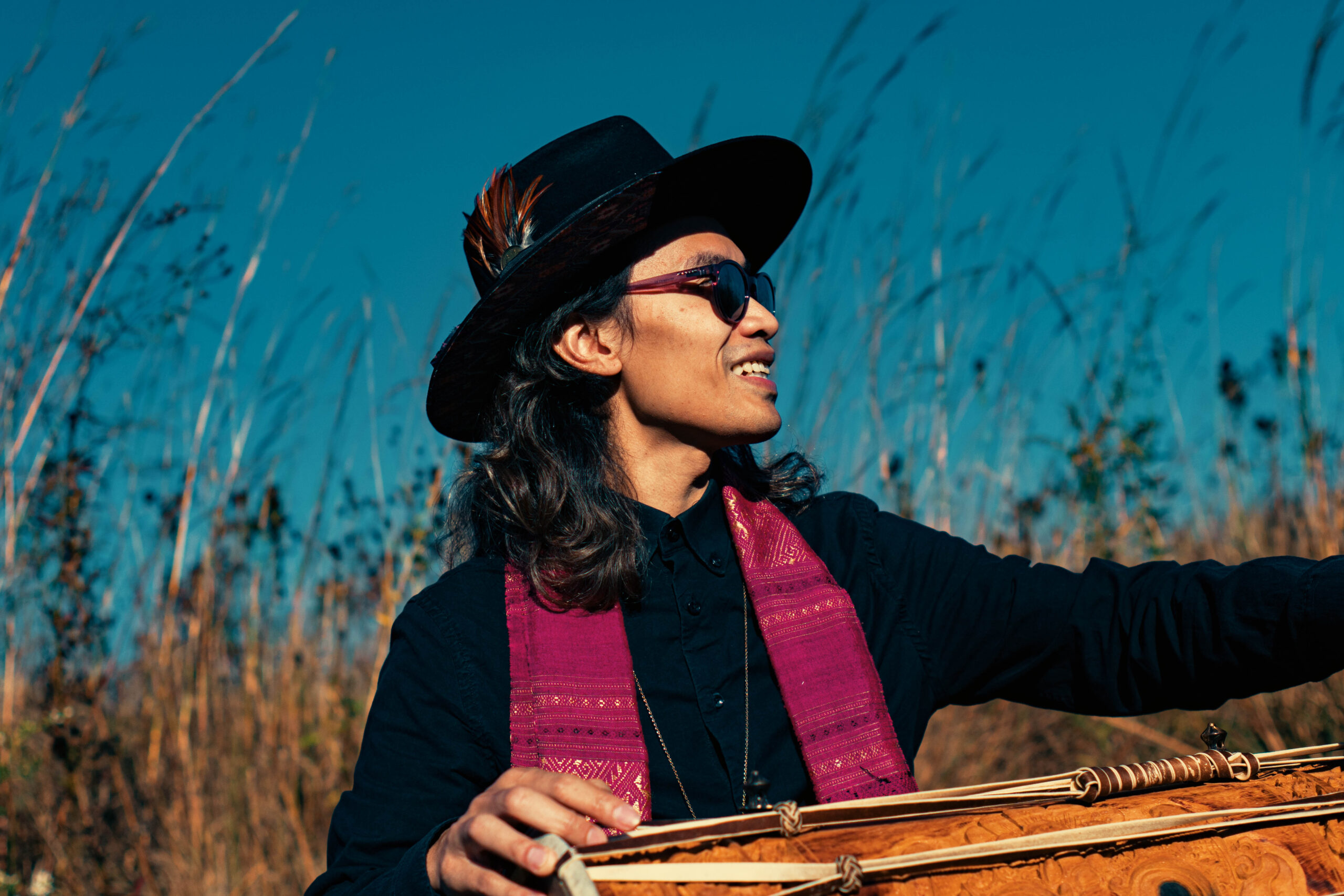 A person looking off to the side. They are wearing a hat and sunglasses, and their hand is resting on a traditional percussive instrument.