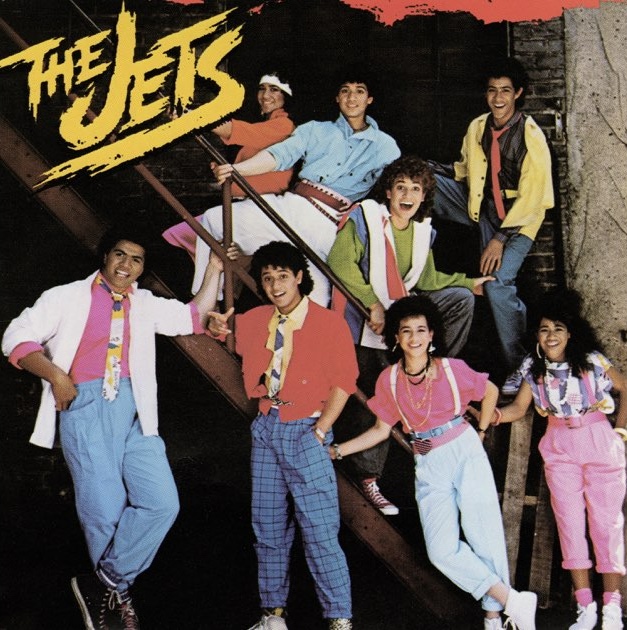 A music album with text that reads "The Jets" over a photo of eight people smiling and posing by a staircase. They are all wearing colorful clothing