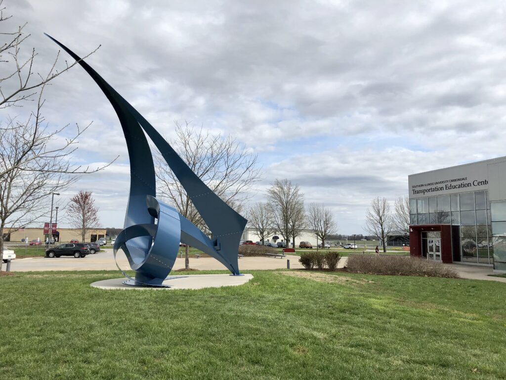 An abstract blue-colored metal sculpture located outside a building with glass windows.