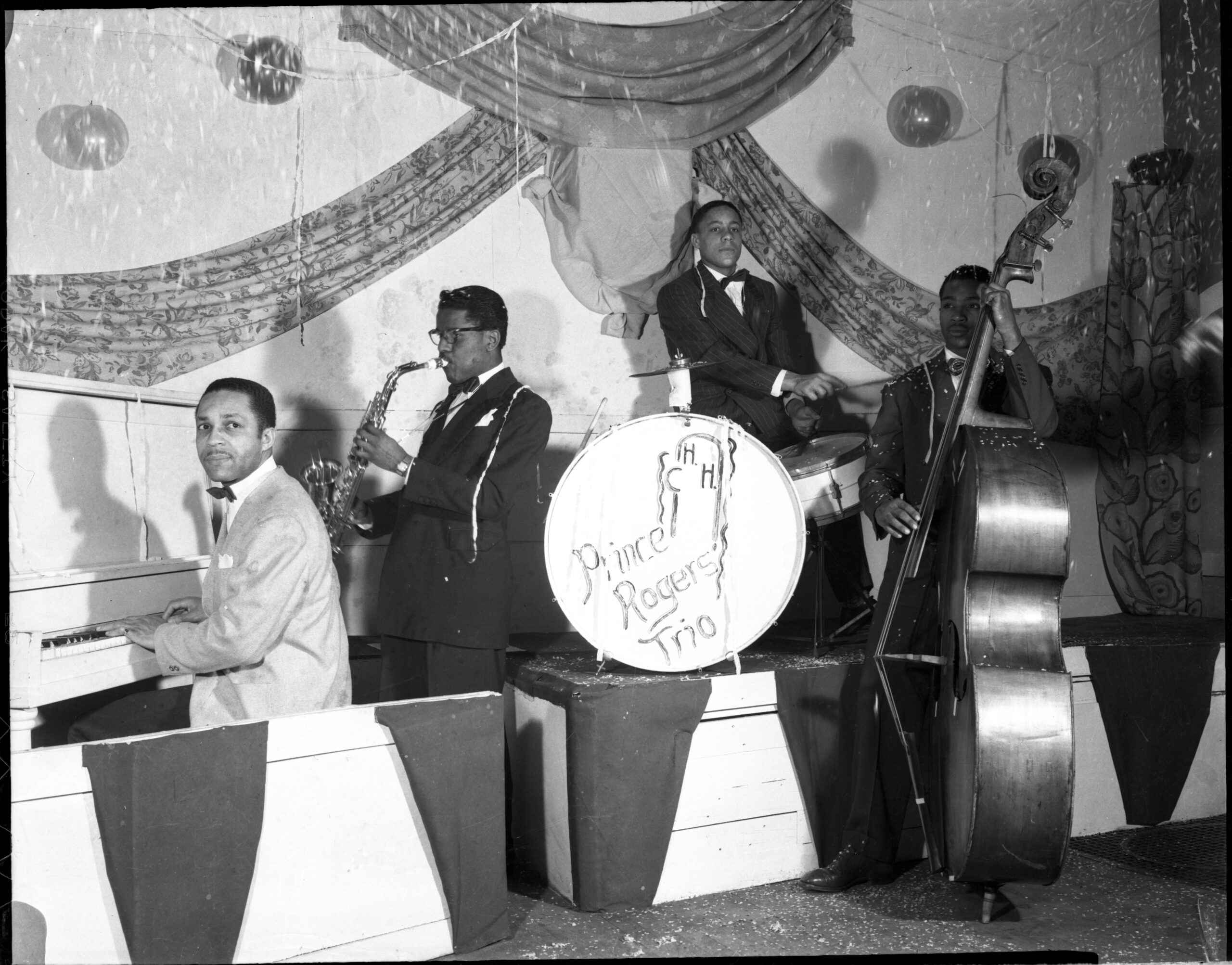 An archival black and white photo of a four-member band playing different instruments from an upright piano to an upright bass. The drums have faint text that reads "Prince Rogers Trio"