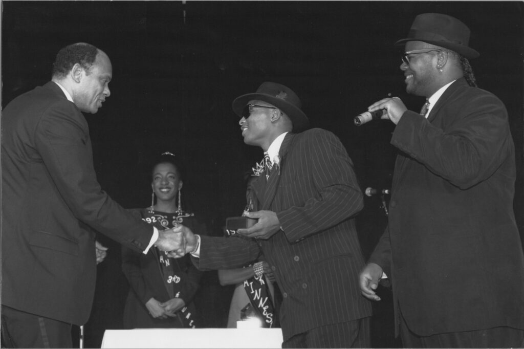 A black and white archival photo of two people receiving an award. Two people are shaking hands while a third stands by them smiling and holding a microphone