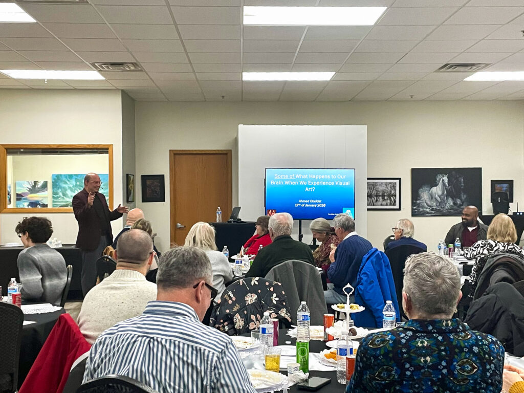 People sitting at tables with drinks and plates of food as a speaker in the front of the room presents to them.