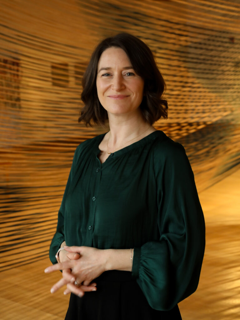 Headshot of a woman with short dark hair, wearing a dark green shirt, in front of a large artwork.