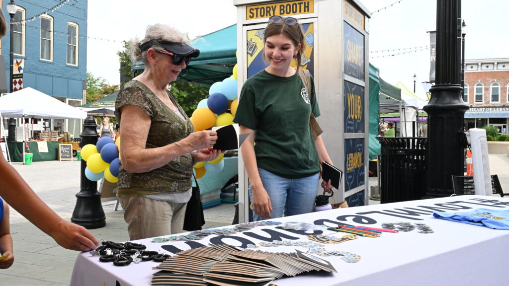 Two people standing next to each other smile as they look at merch on a table at an outdoor event