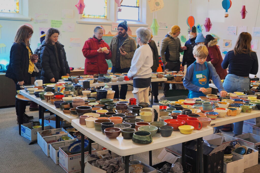 A room of people looking at bowls set up on white tables in a square.