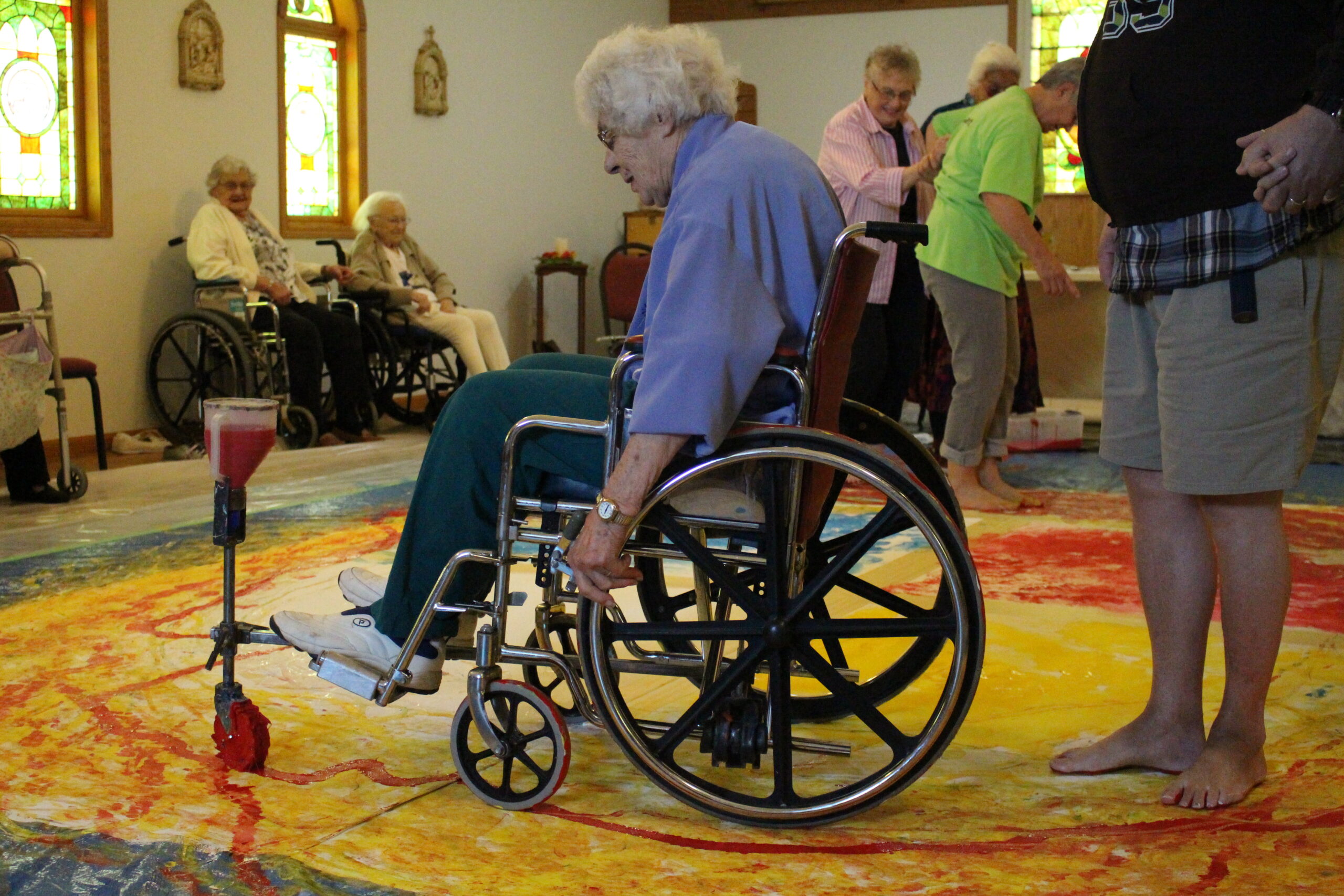 An elderly person sitting in a wheelchair with a mobile painting device attachment. They are using the chair to paint on a large canvas on the floor. Other elderly people behind them are painting on the canvas with their feet.