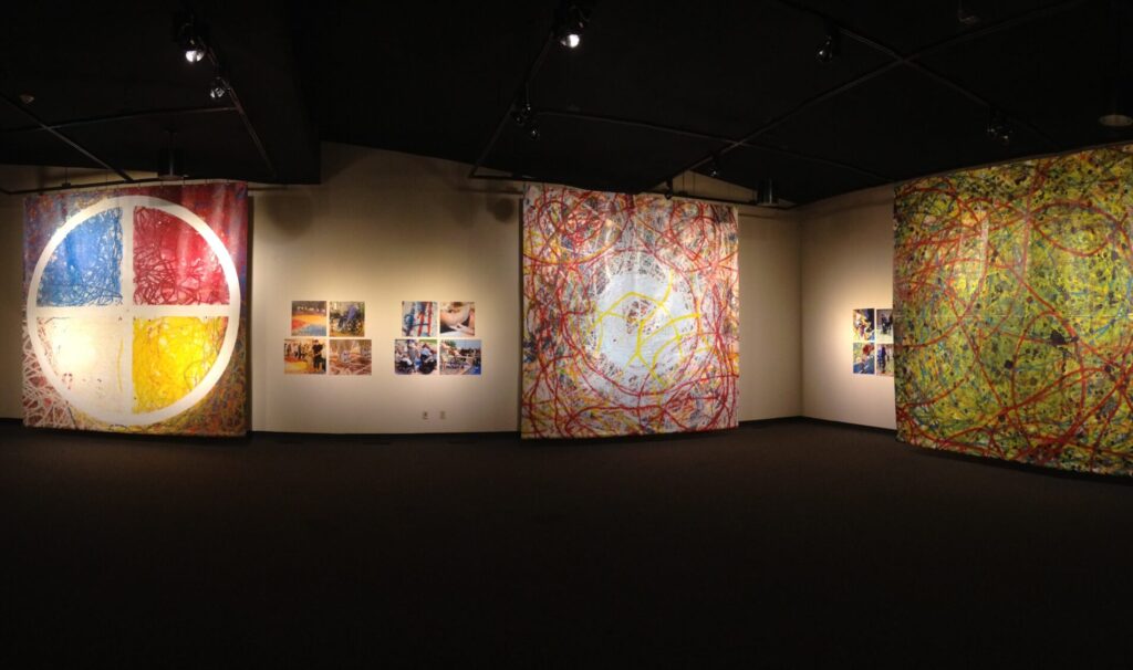 Three large floor-to-ceiling paintings hanging in a gallery space. The wall space between the paintings have photo works displayed.
