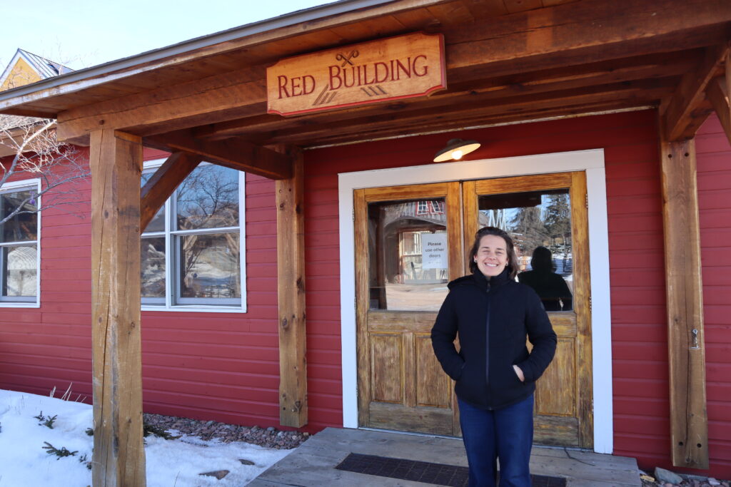 A woman with light skin smiles outside a red building.