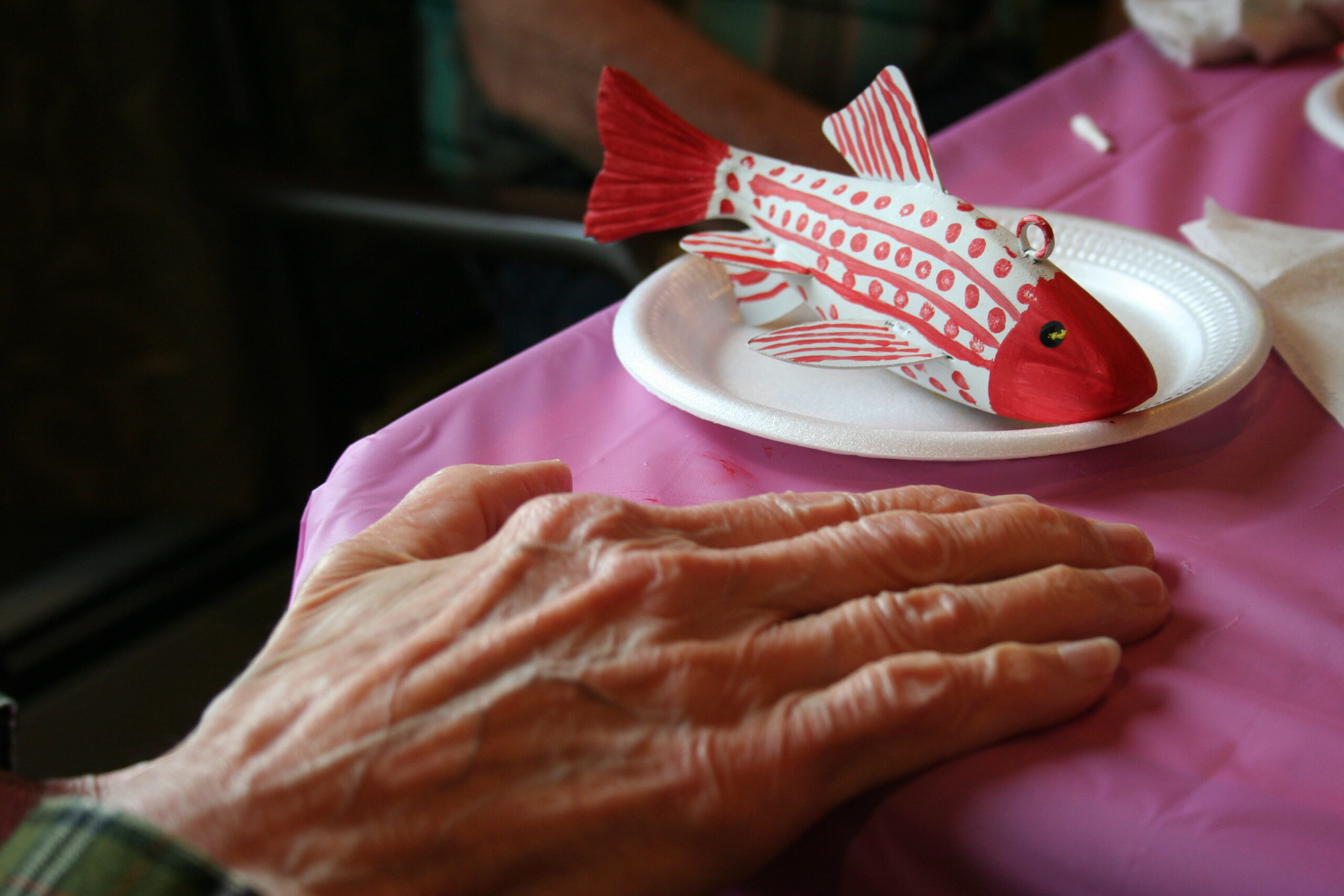 A freshly painted red and white fish decoy sitting on a paper plate. There's a hand of an elderly person resting by it.