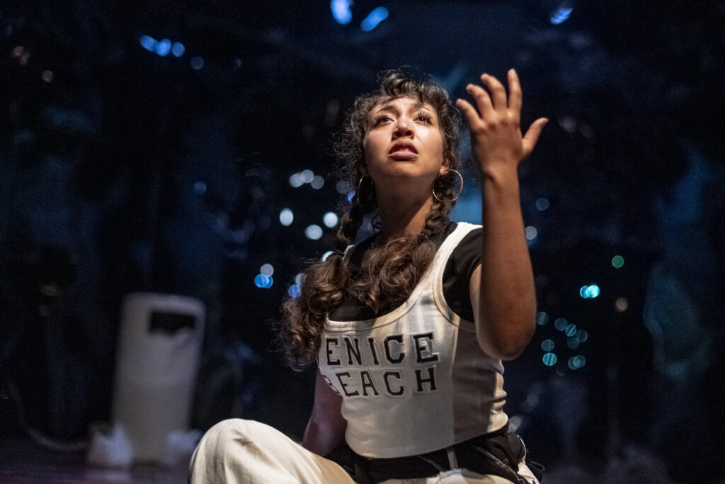 A performer sits on a darkened stage, gazing upward with one hand raised and fingers spread, wearing a "Venice Beach" tank top, hoop earrings, and braided hair. Blue bokeh lights glow in the background.
