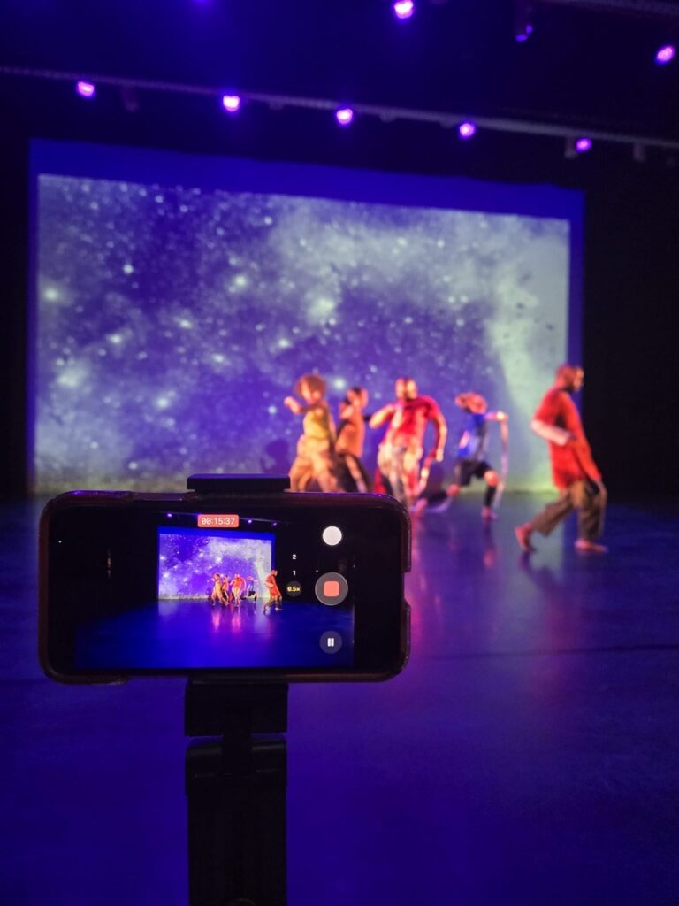 A smartphone on a tripod records a live dance performance in the foreground, while behind it, a group of dancers in colorful costumes move across a blue-lit stage in front of a large projection of a starry night sky.