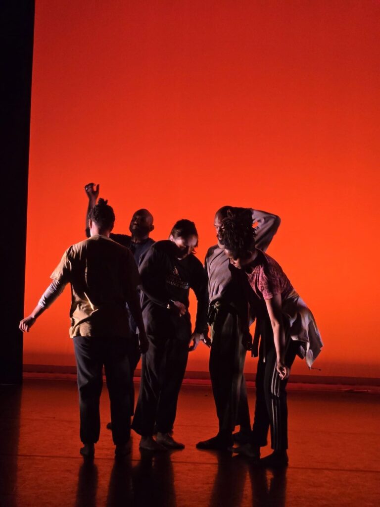 Four dancers cluster together on a dimly lit stage, their bodies in varied poses — one arm raised, another bent forward — silhouetted against a vivid red-orange backdrop.