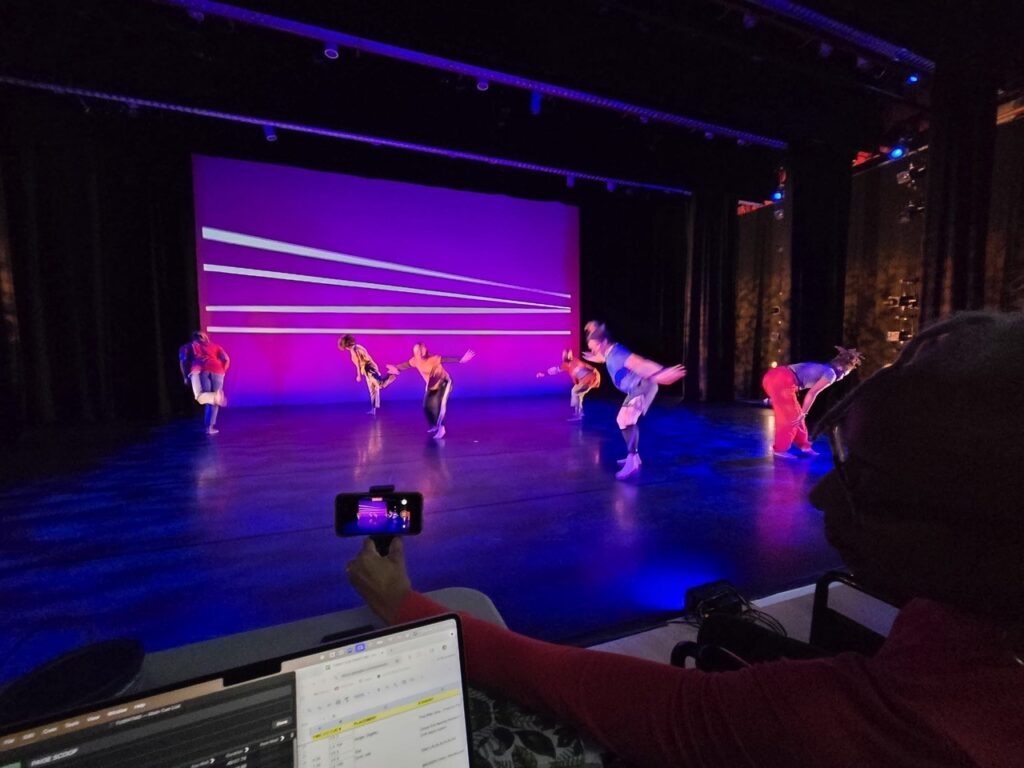 Viewed from the audience, a person holds a smartphone to record a dance performance while a laptop sits open in the foreground. On stage, six dancers in colorful costumes move dynamically under purple and blue lighting in front of a backdrop with white diagonal lines.