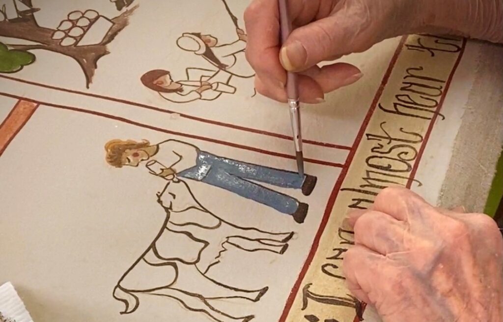 A elderly person using a paintbrush to fill in color in an outline drawing depicting a person feeding a cow