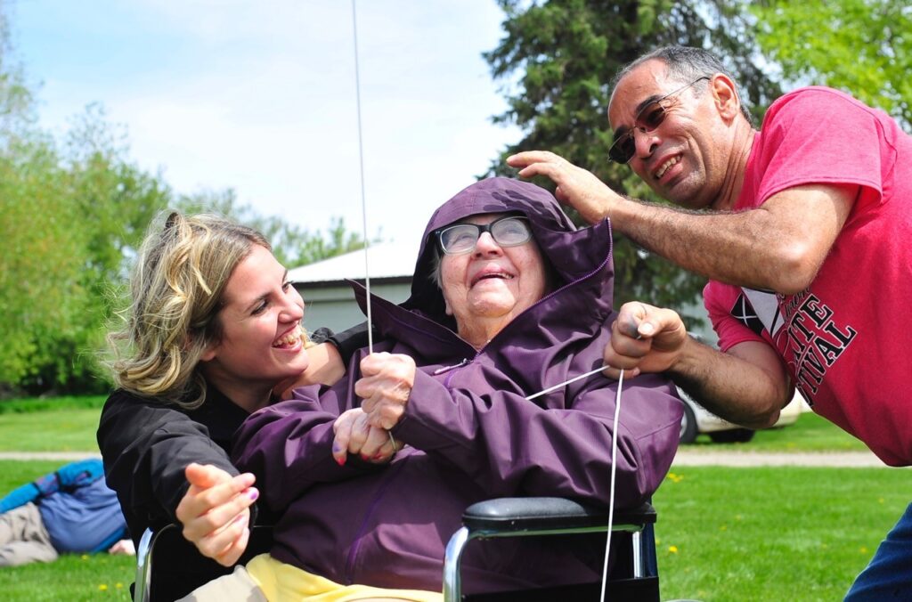 A person sitting in a chair holding a kite line with the assistance of two people standing next to them.