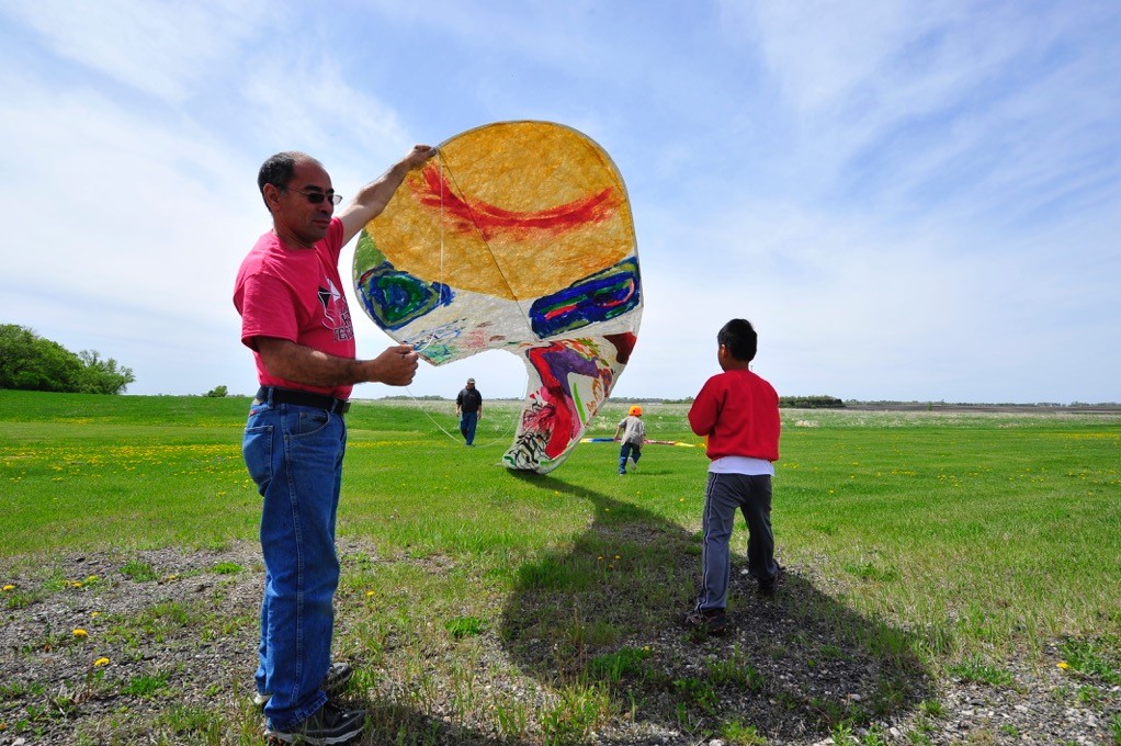 An adult holding a large colorful kite in a big field outdoors while two children run around.