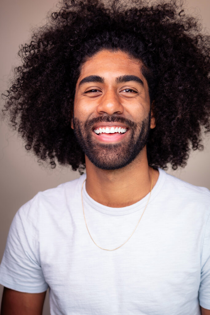 Headshot of a smiling man with brown skin tone and big curly hair, wearing a light blue shirt.