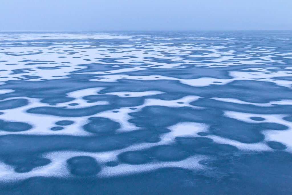 A frozen lake with a horizon line at the far end of the photo. There is snow on the icy lake and it's made various abstract patterns on the surface