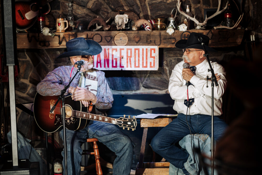 Two people wearing cowboy hats, button-up shirts, and blue jeans sit on stools on a stage. One of them is holding an acoustic guitar, while the other holds a microphone.