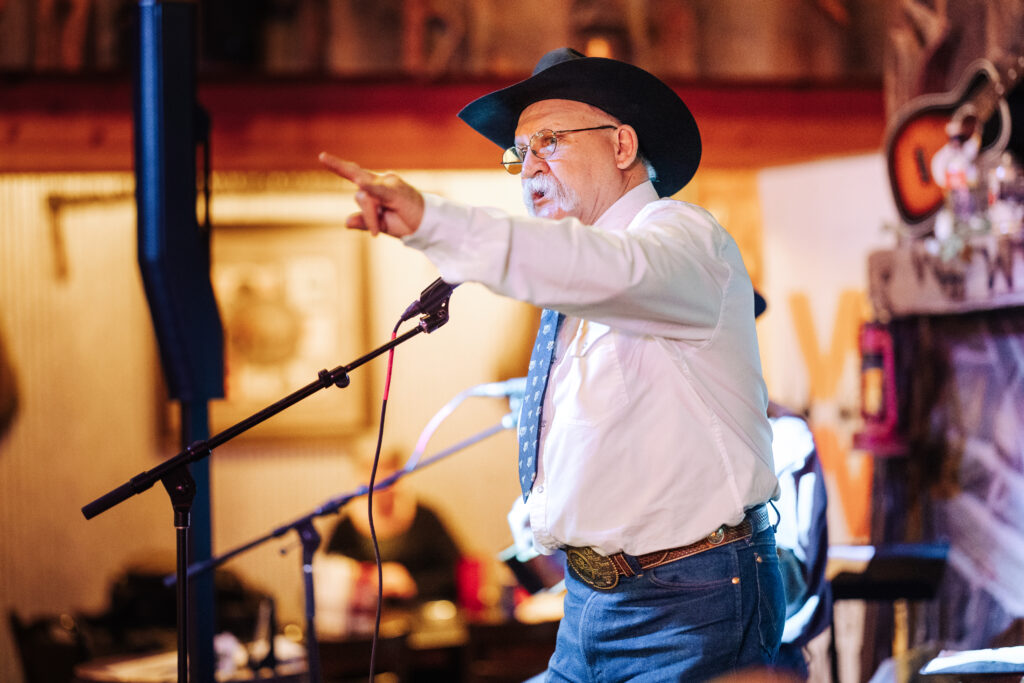A person speaking into a microphone, with one arm raised. They are holding up their hand to make "horns" with their pointer and pinky fingers.