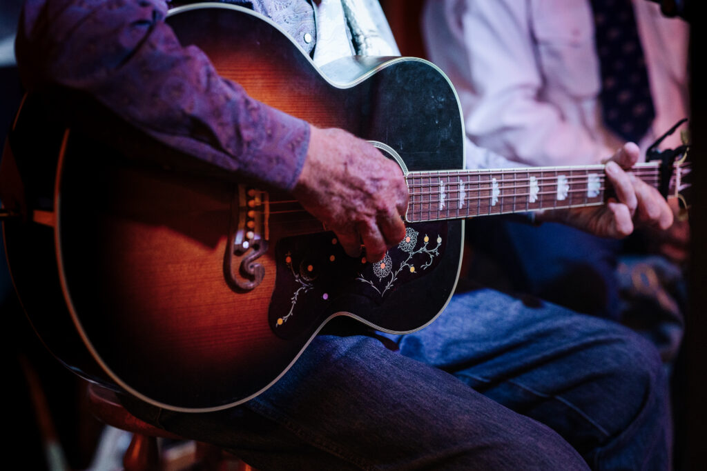 A person strumming an acoustic guitar. They are sitting and resting the guitar on a leg. Only their arms and legs are in view in this photo.