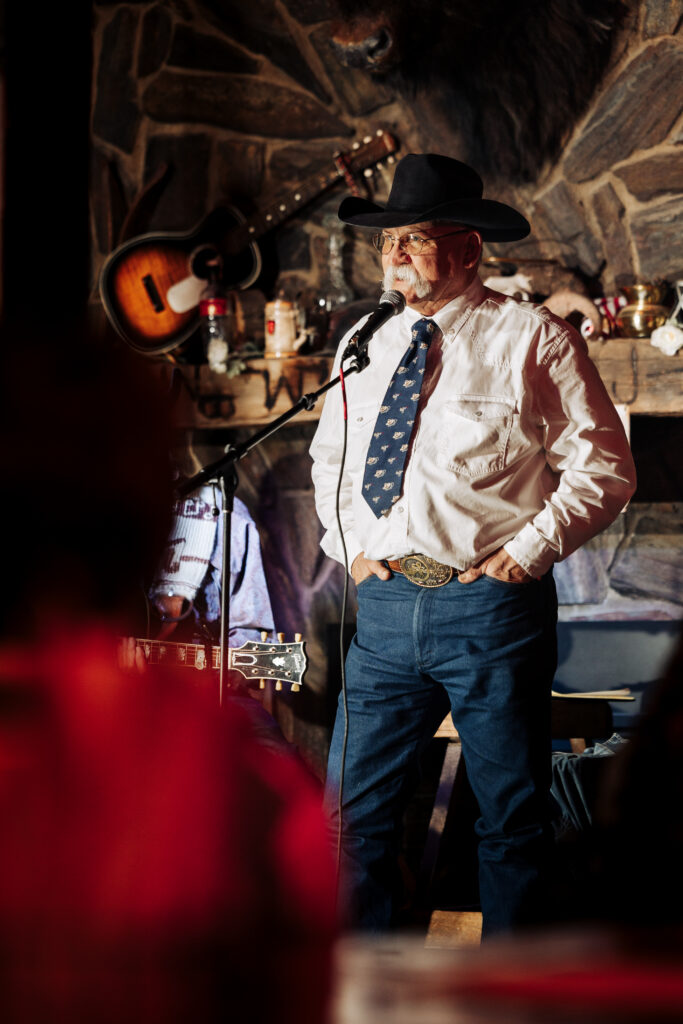 A person standing and speaking into a microphone. They have a pronounced mustache and are wearing a dark cowboy hat, a white long sleeve shirt with a blue necktie, blue jeans and a belt with a big buckle.