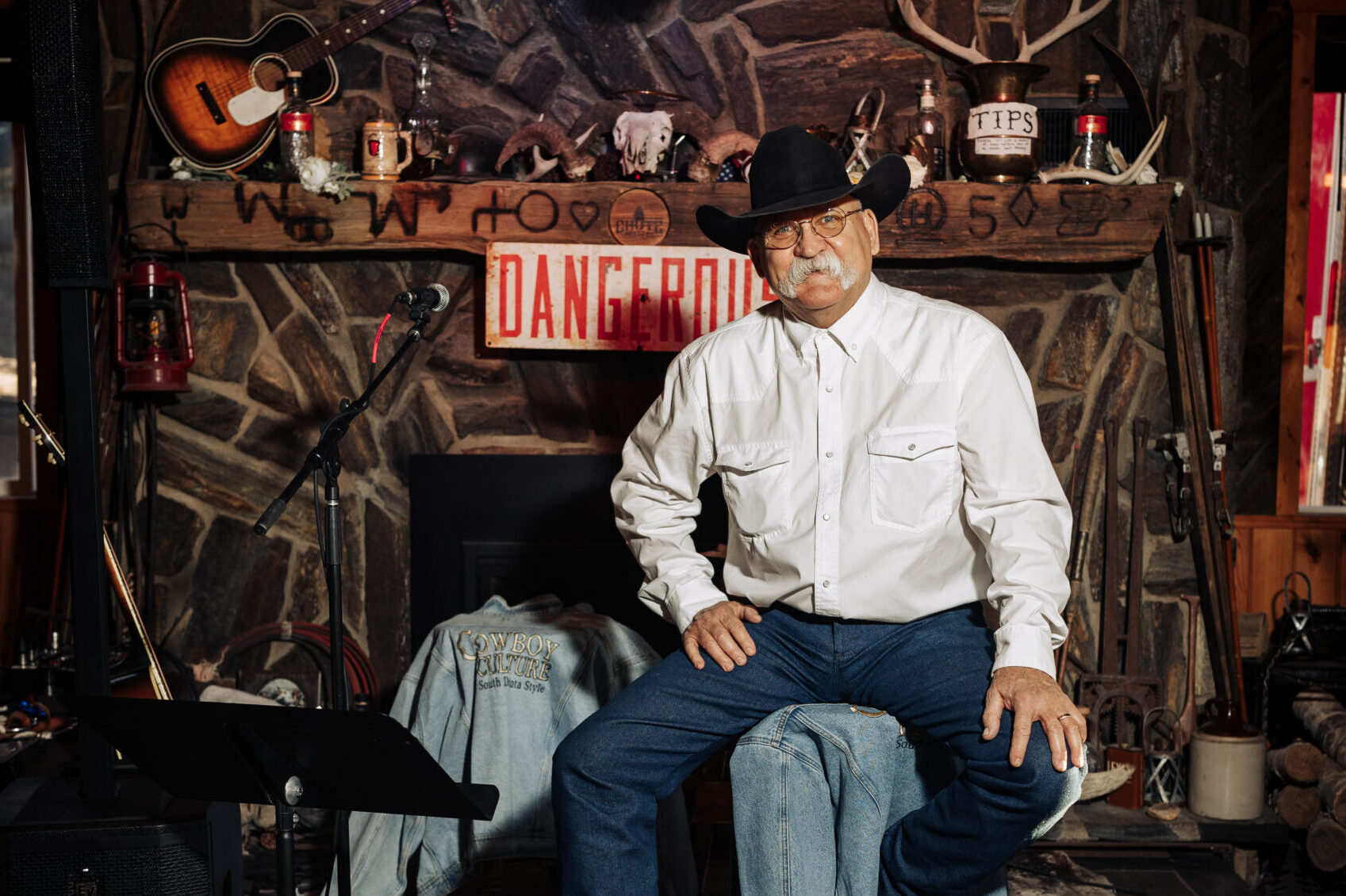 A person with a white mustache sitting on a tall stool and smiling. They are wearing a cowboy hat, thin-framed glasses, a white button-up shirt, and blue jeans.