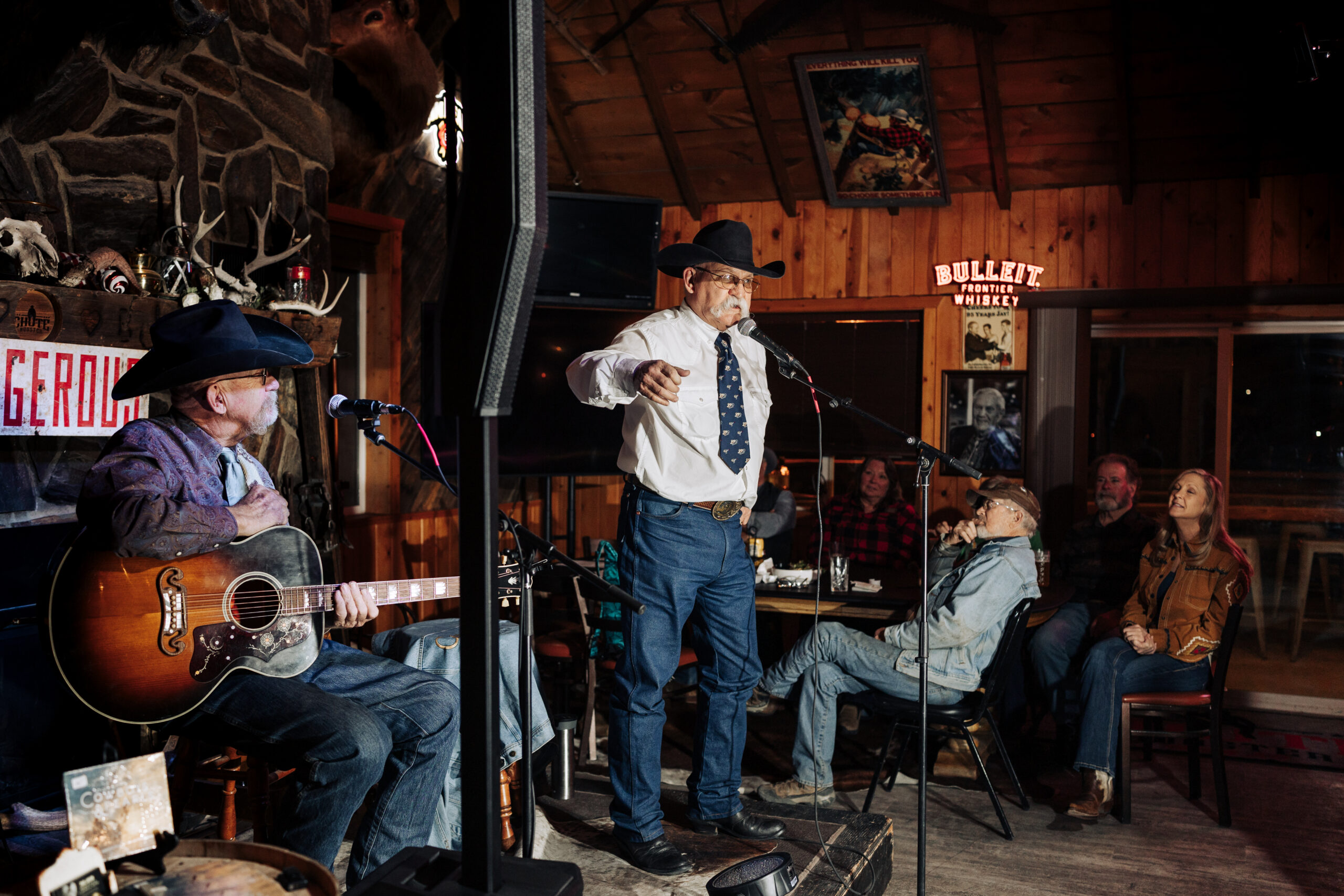 Two people performing on stage as a seated audience looks on. Both of them are wearing cowboy hats, button-up shirts, and blue jeans.