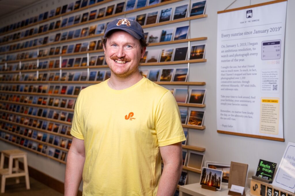A smiling person with light skin tone wearing a yellow tshirt and a navy blue baseball cap.