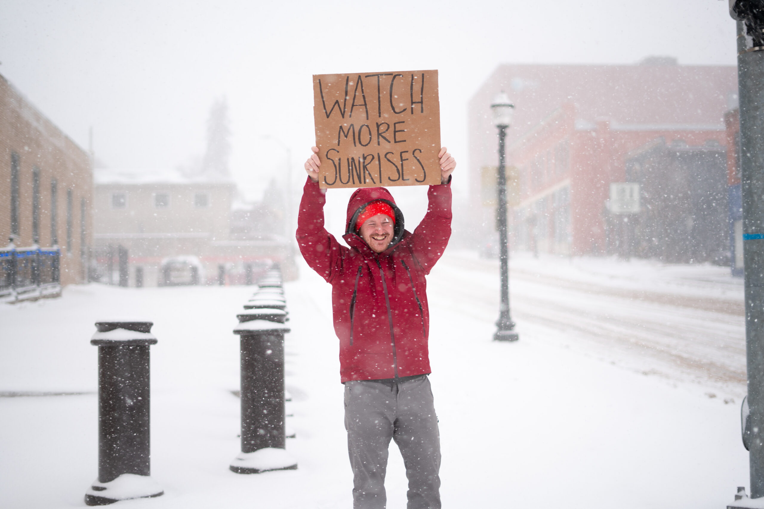 A person wearing a red winter coat and bright hat standing in the street as snow falls. They are holding up a cardboard sign with sharpie text that reads "watch more sunrises"