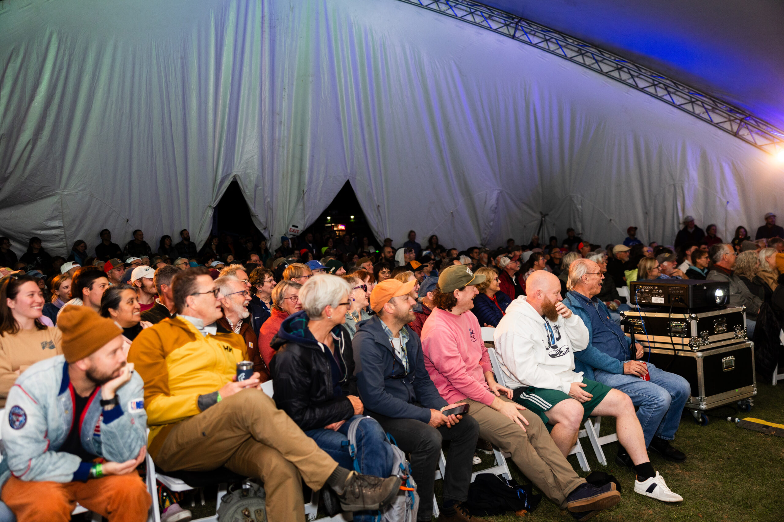 A large audience of people sitting in chairs, all facing one direction under a large tent-like structure.