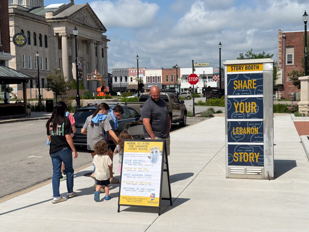 A family of adults and young people walking on a sidewalk as they look at a refurbished phone booth and signage next to it