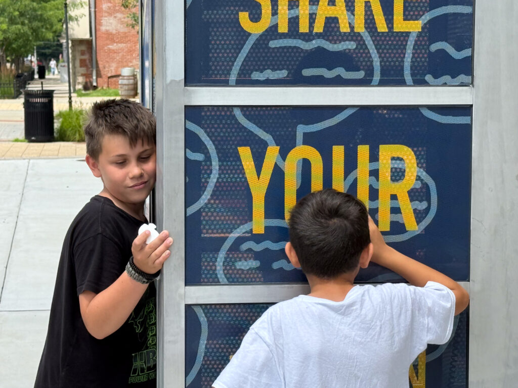 Two young people pressing their ear and face against a refurbished phone booth