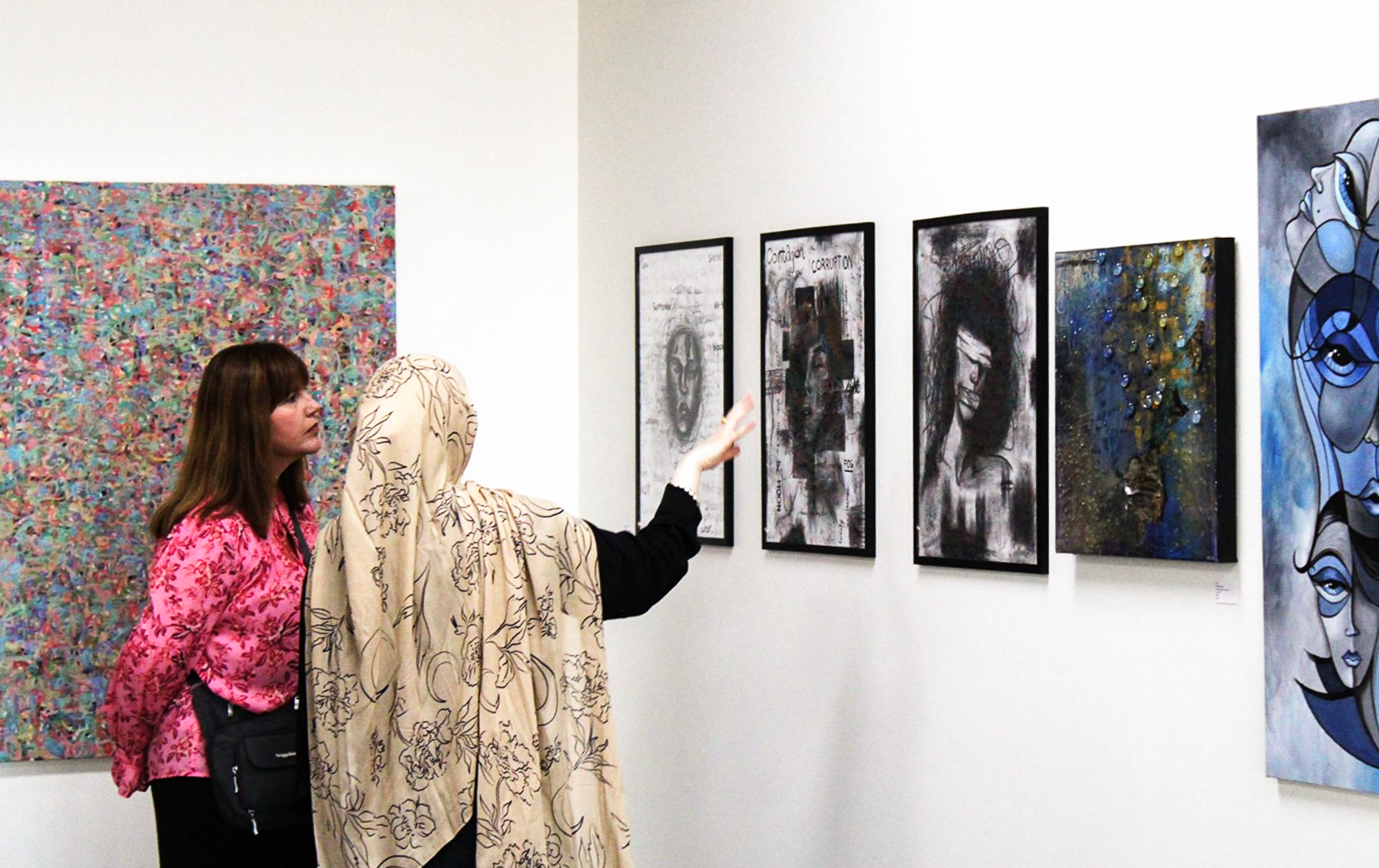 Two people standing together by a wall of paintings. One person is gesturing towards a painting as the other looks on.