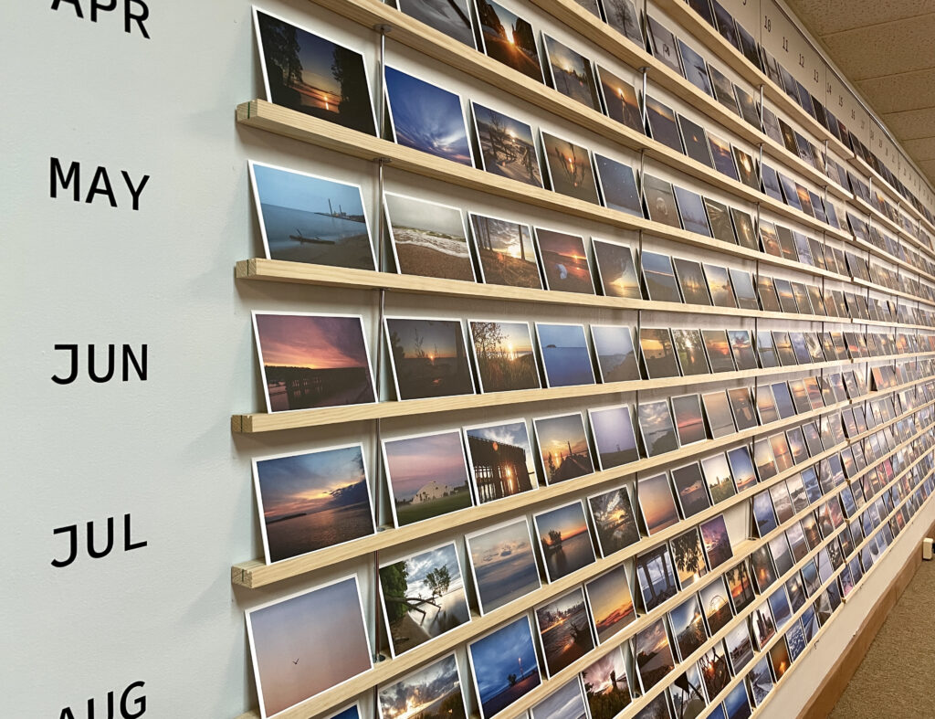 A wall with shelves from floor to ceiling with photo prints of sunrises. There are months and dates listed on the side and top of this wall of shelves.