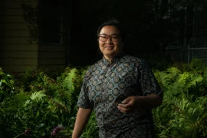 Valerie Oliveiro, a person with medium skin tone, short dark hair, and round tortoiseshell glasses smiles warmly at the camera, wearing a patterned short-sleeve button-up shirt, standing in front of lush green ferns outdoors.